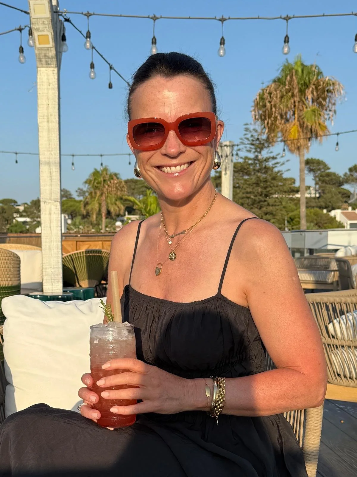 A woman with dark hair in a bun, wearing large red sunglasses, gold jewelry, and a black spaghetti strap dress, smiling and holding a cocktail with a straw and garnish, sitting outdoors at a sunny patio with string lights and palm trees in the background.