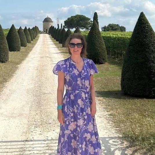 Woman in a purple floral dress standing on a gravel path lined with trimmed cone-shaped bushes in a garden, with a domed structure and trees in the background on a sunny day.
