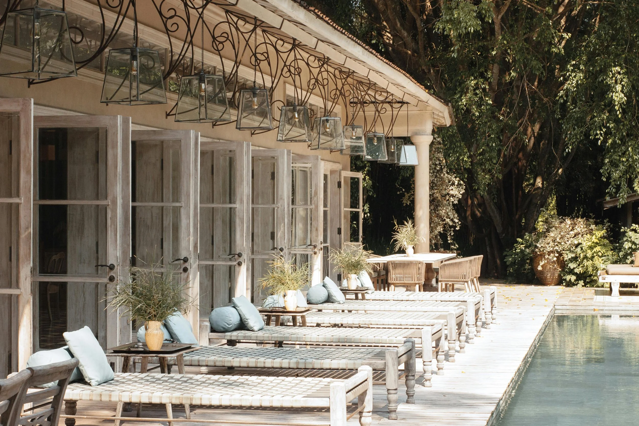 Outdoor seating area with wooden benches and chairs, cushions, small tables with vases, a poolside edge, and trees in the background. Hanging lanterns are above the seating area.