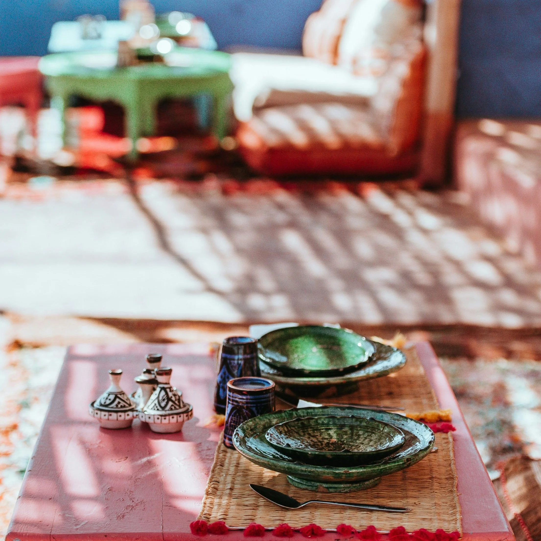 A row of colorful ceramic plates and bowls on a pink table with a woven placemat and a black spoon. In the background, there are small decorative ceramic containers, possibly for spices or condiments.