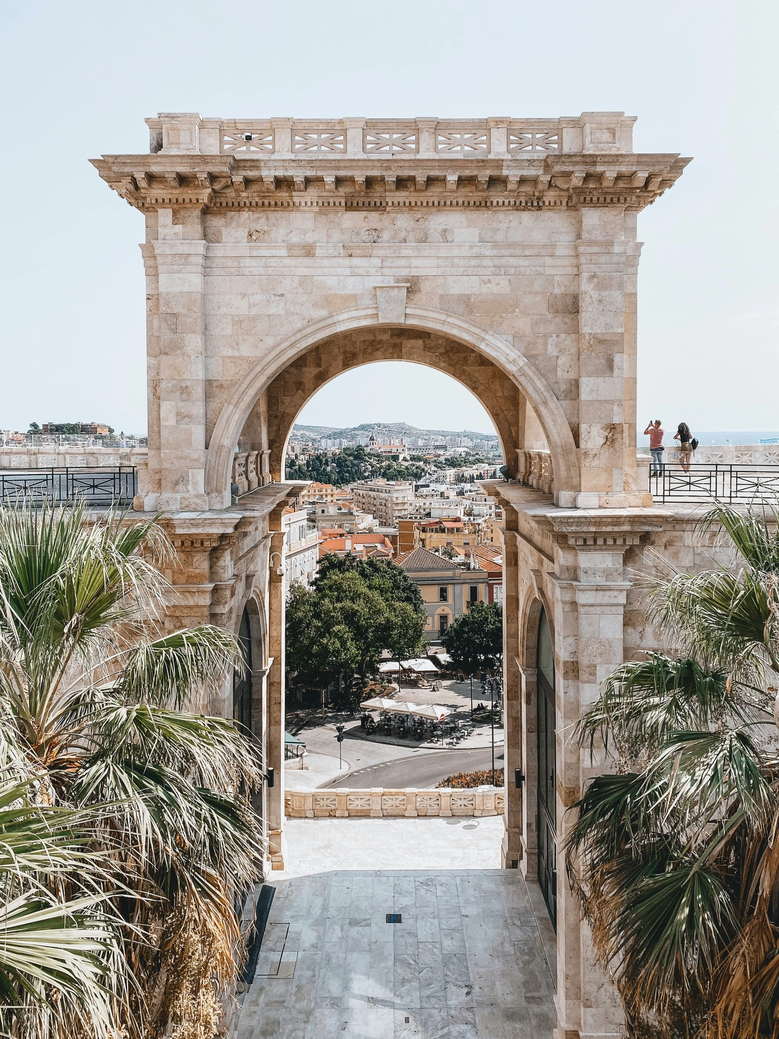 Architectural archway with cityscape view, palm trees, and people taking photos in the distance.