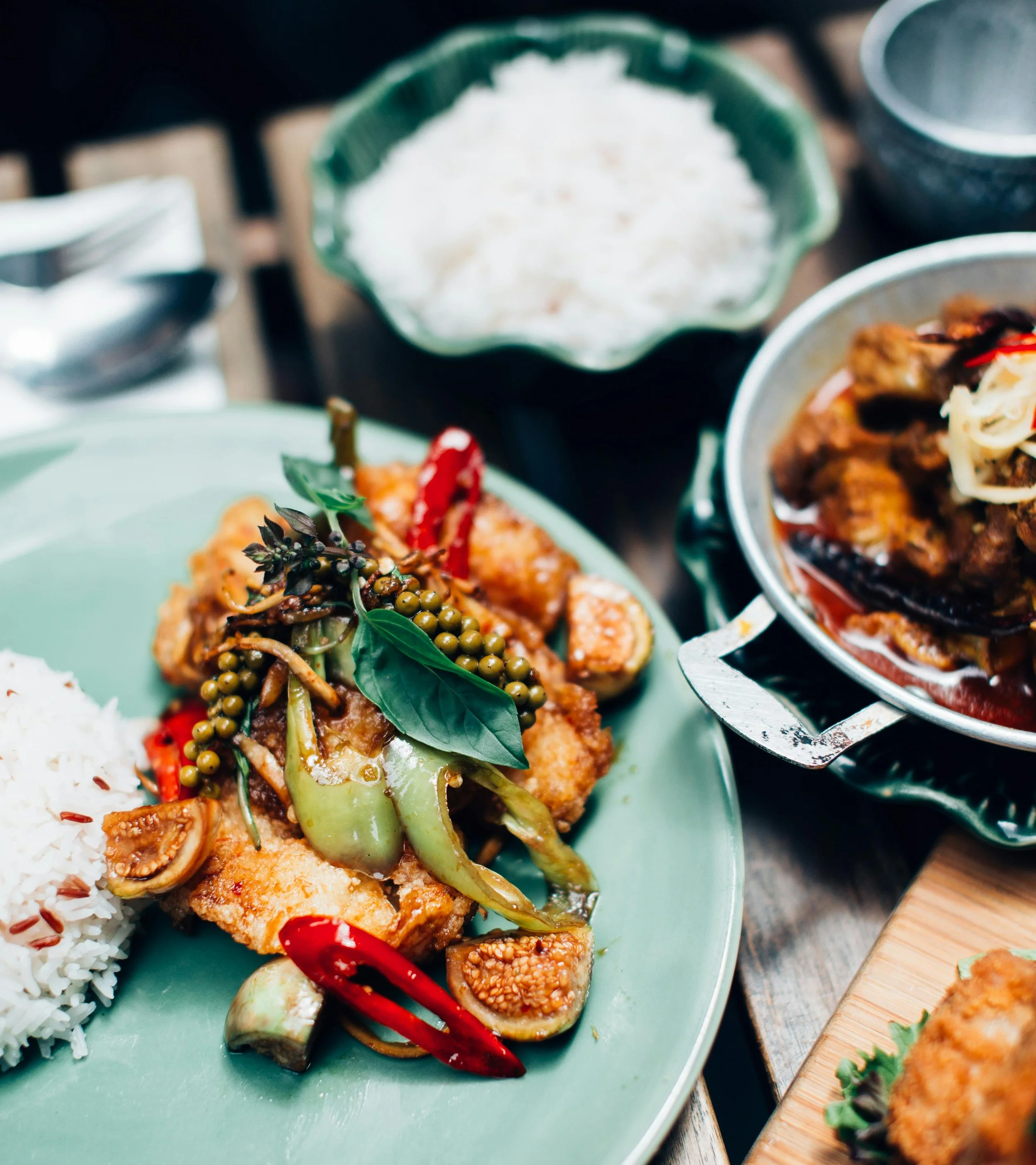 A plate of Asian-style fried fish topped with vegetables and herbs, with cooked white rice on the side, set on a table with a spoon and bowls nearby.