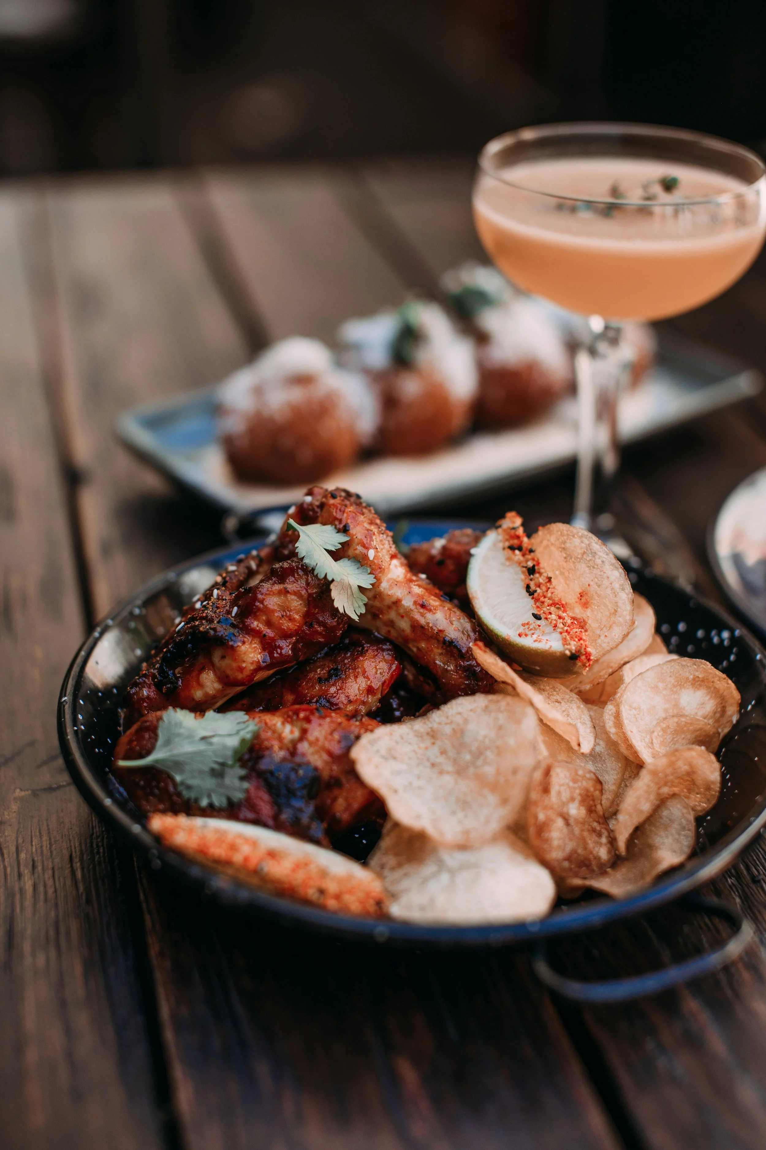 A black bowl of buffalo chicken wings garnished with cilantro, with a sliced lime on the side, surrounded by potato chips, served on a dark wooden table.