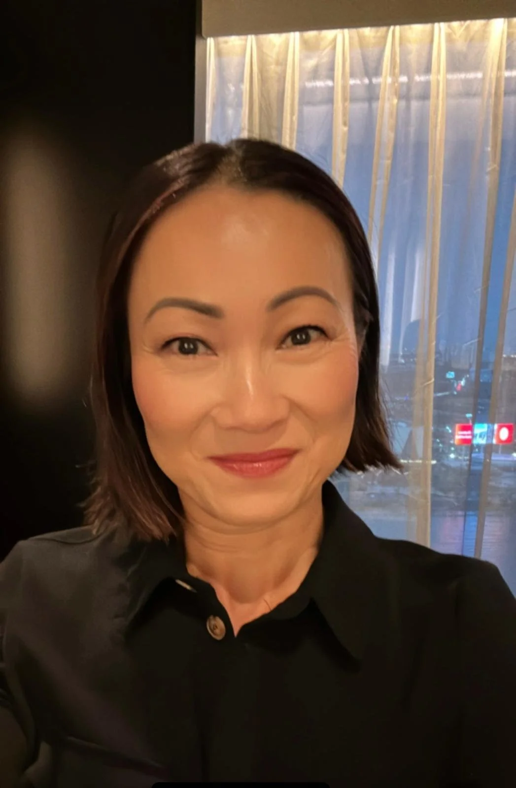 A woman with short dark hair, wearing a black shirt, smiling, sitting indoors near a window with sheer curtains, city lights visible outside.