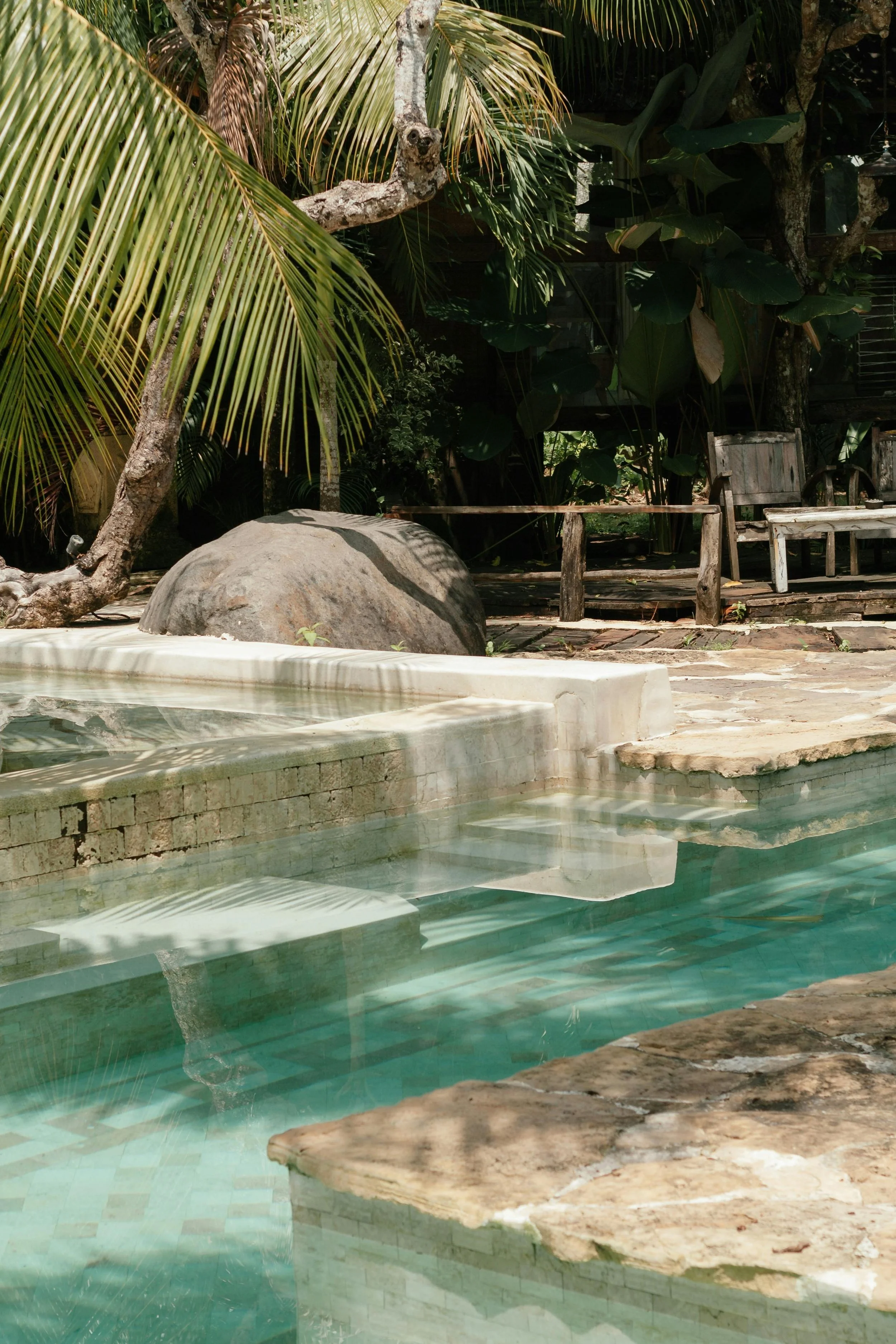 A small outdoor swimming pool with water reflecting surrounding palm and other tropical plants. There are two worn wooden chairs and a wooden deck nearby. Large rocks decorate the pool area.