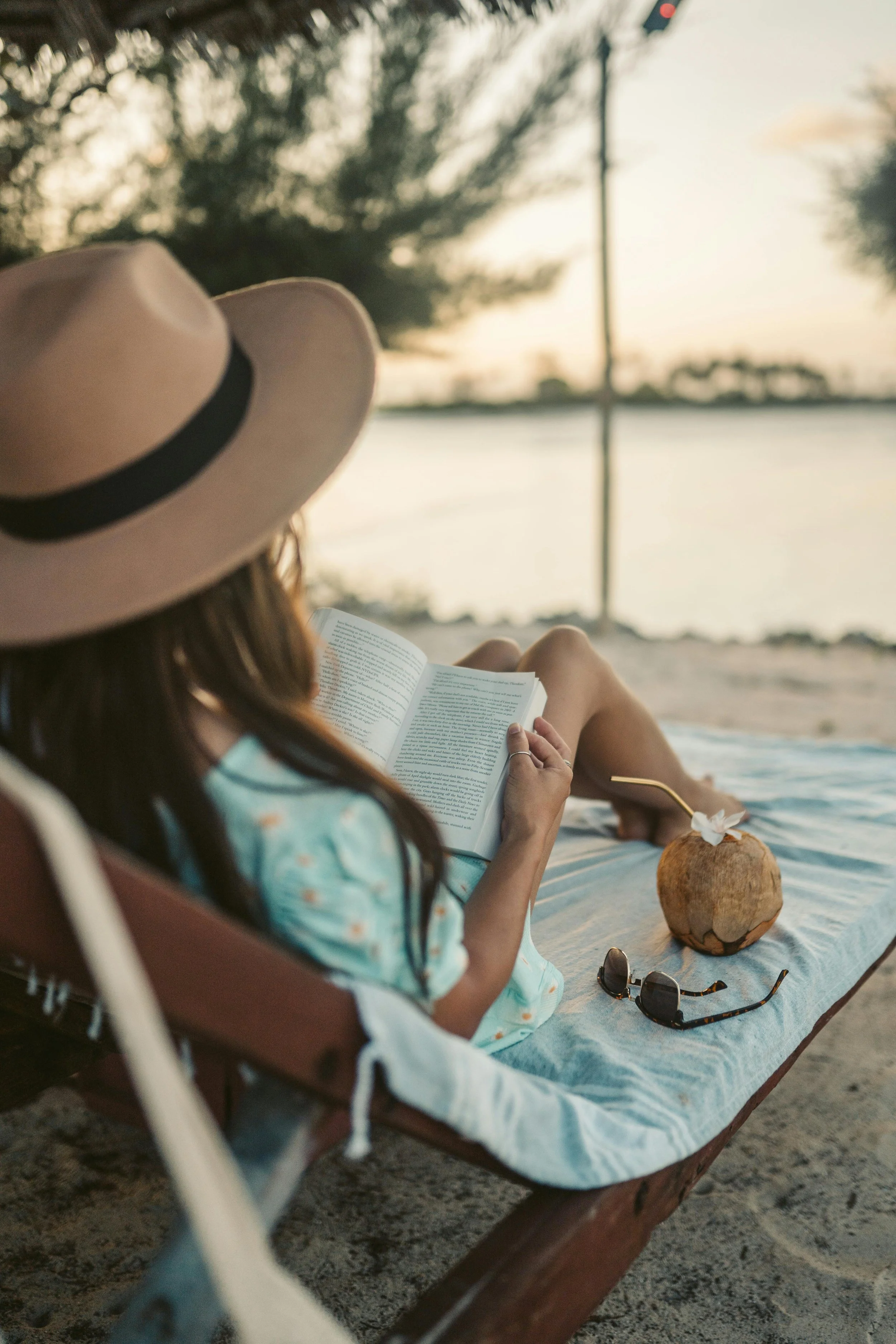 A woman lying on a lounge chair at the beach, reading a book at sunset. She has a large coconut drink with a straw and a flower on it beside her, and sunglasses and a patterned headband nearby. She is wearing a hat and a blue dress with a floral pattern.