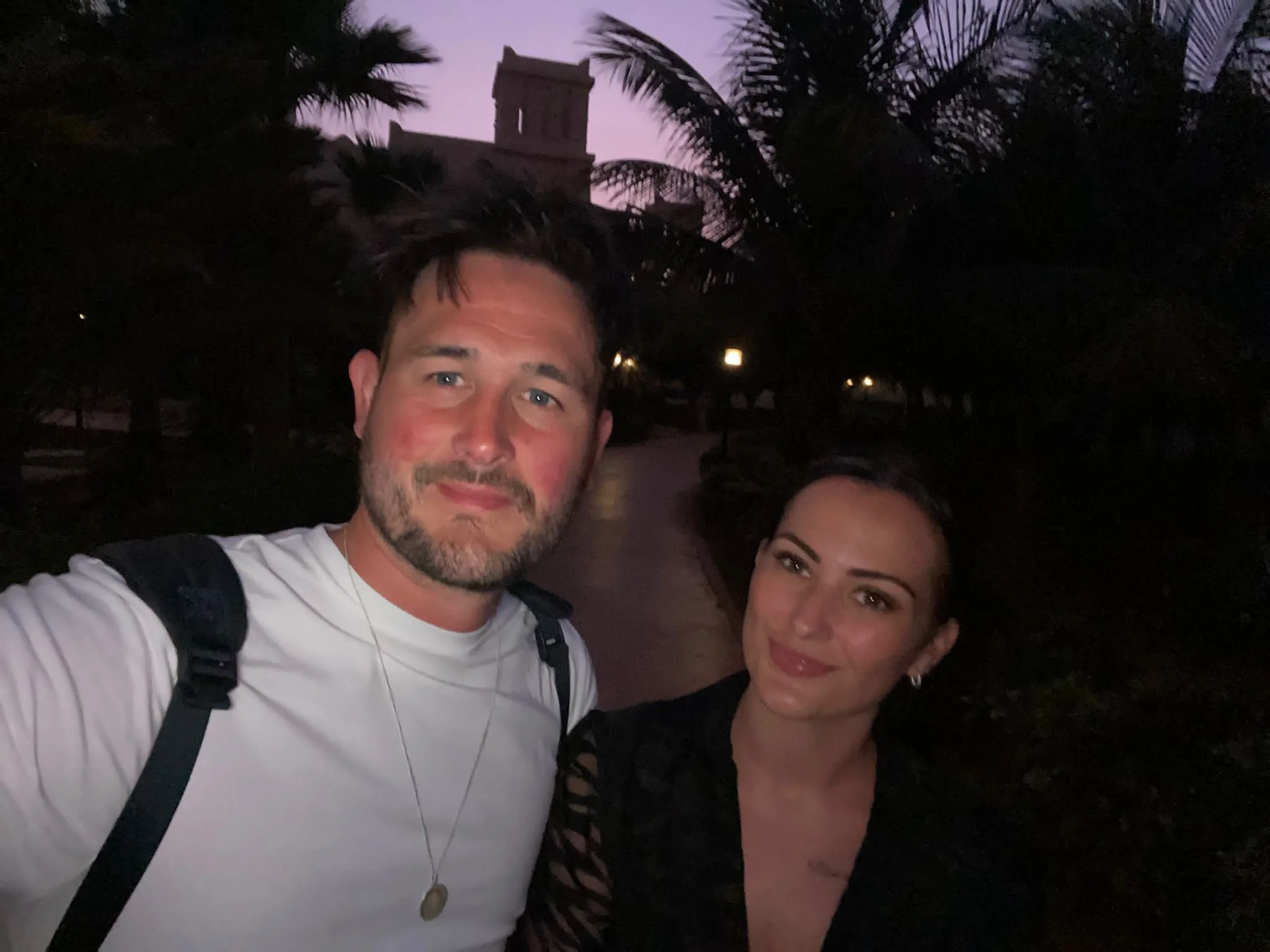 A man with short dark hair and a beard, wearing a white shirt and a backpack, standing with a woman with long dark hair, in front of palm trees during dusk or evening.