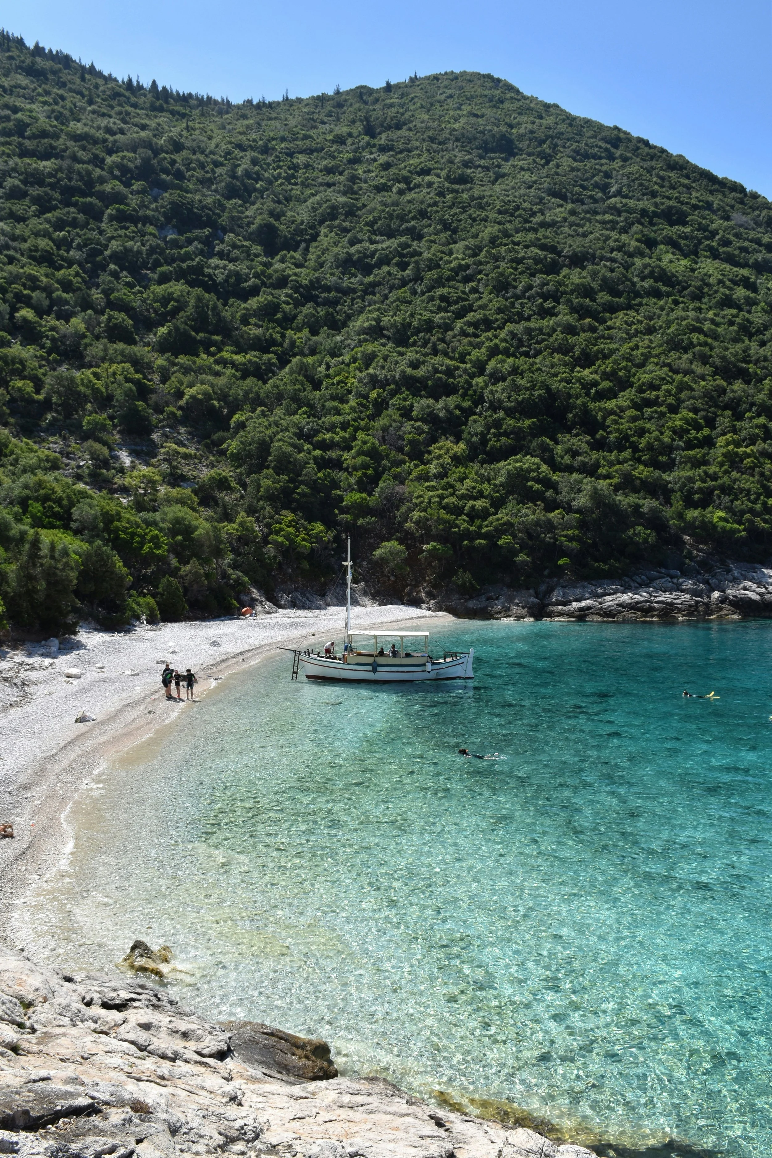 A small boat anchored near a pebble beach with clear turquoise water, surrounded by green hills under a clear blue sky.