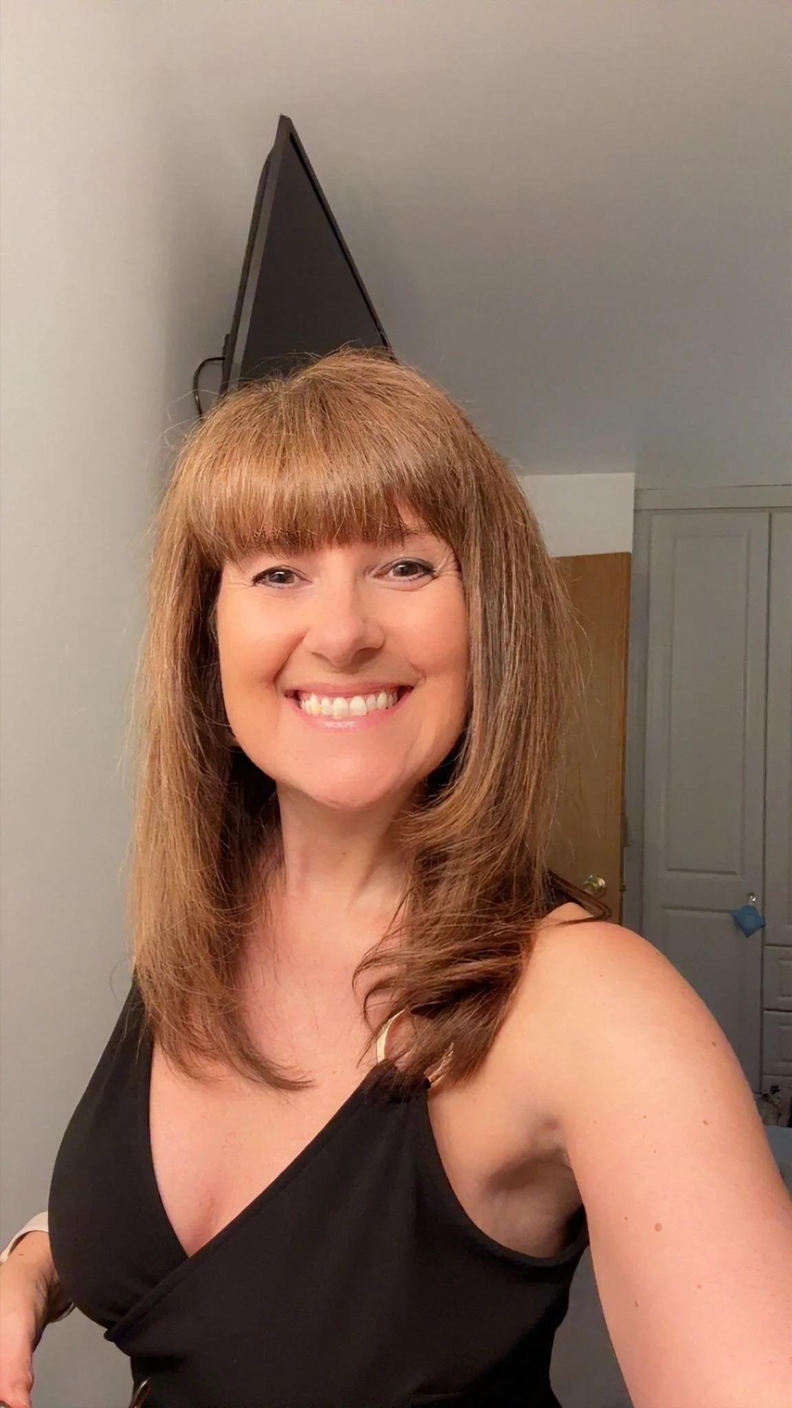 A woman with long, reddish-brown hair and bangs, smiling in a black sleeveless top, taking a selfie in a room with gray walls, a closed closet door, and a small blue object hanging on the door knob.