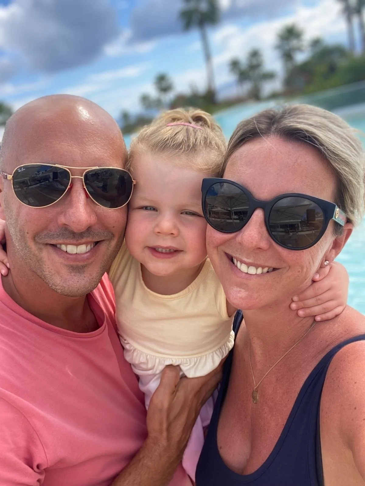 A smiling family of three taking a selfie near a pool with palm trees in the background. The man and woman are wearing sunglasses, and the young girl is between them.