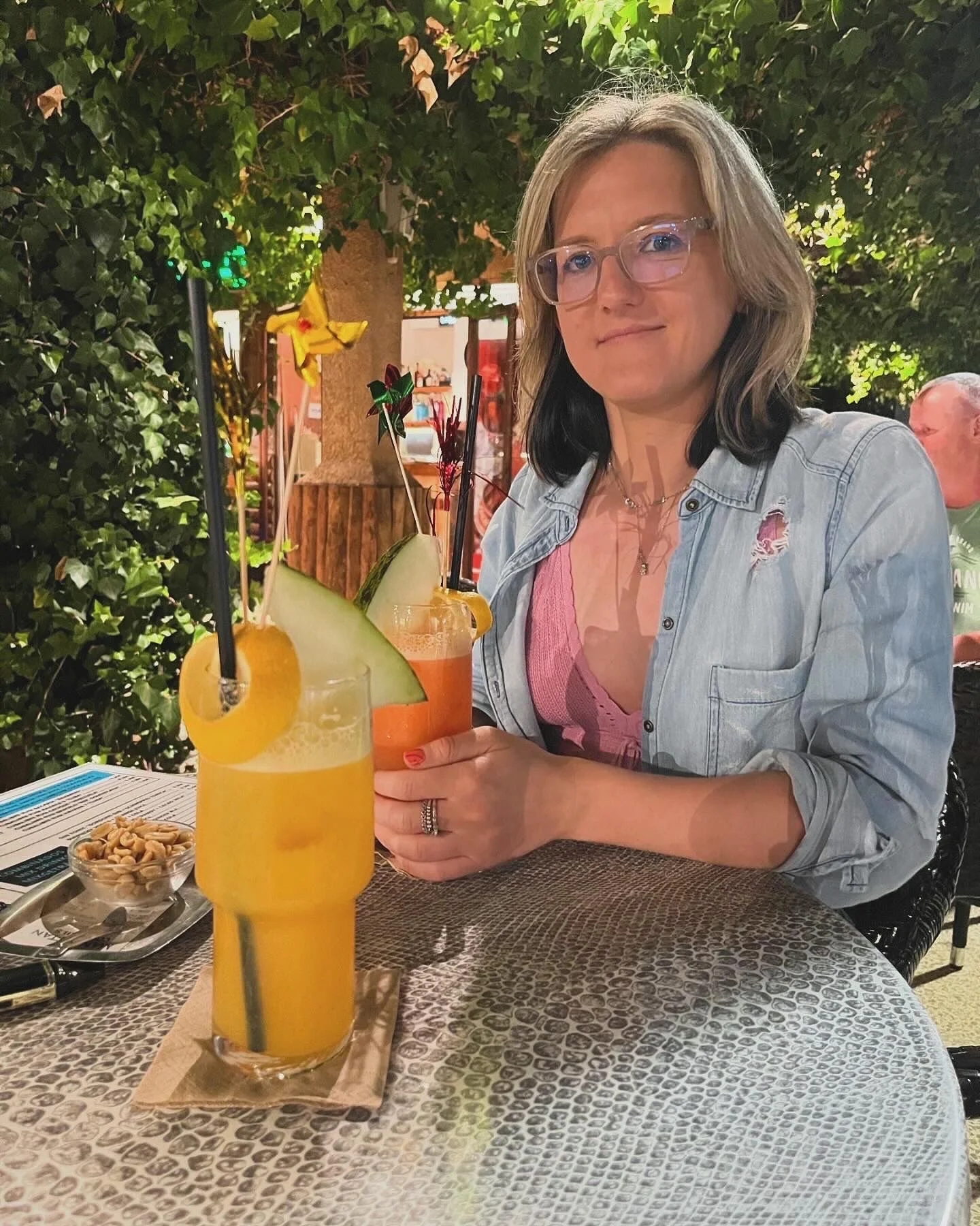 A woman with glasses sitting at an outdoor restaurant table with a colorful drink garnished with fruit and a straw. There is a green hedge in the background and a man sitting behind her.