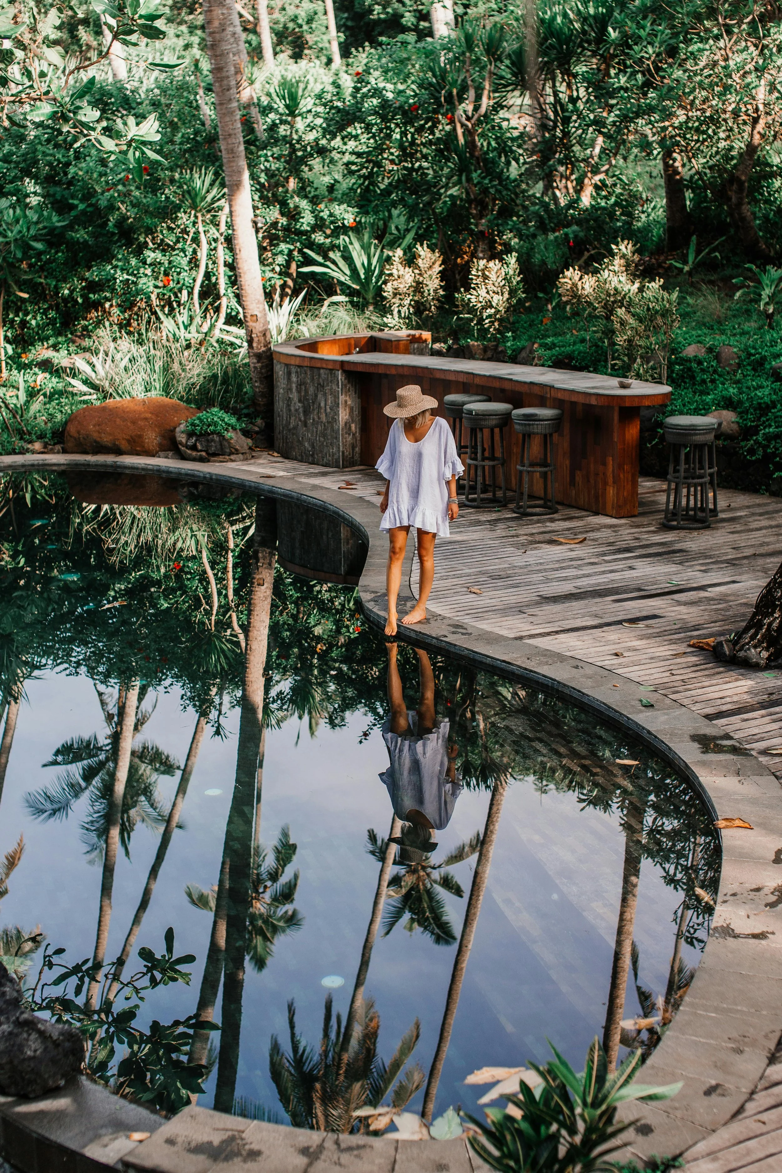 A woman in a white dress and wide-brimmed hat standing at the edge of a small pond, with her reflection visible in the water. The pond is surrounded by lush green tropical plants and trees, with a wooden deck and barstools nearby.