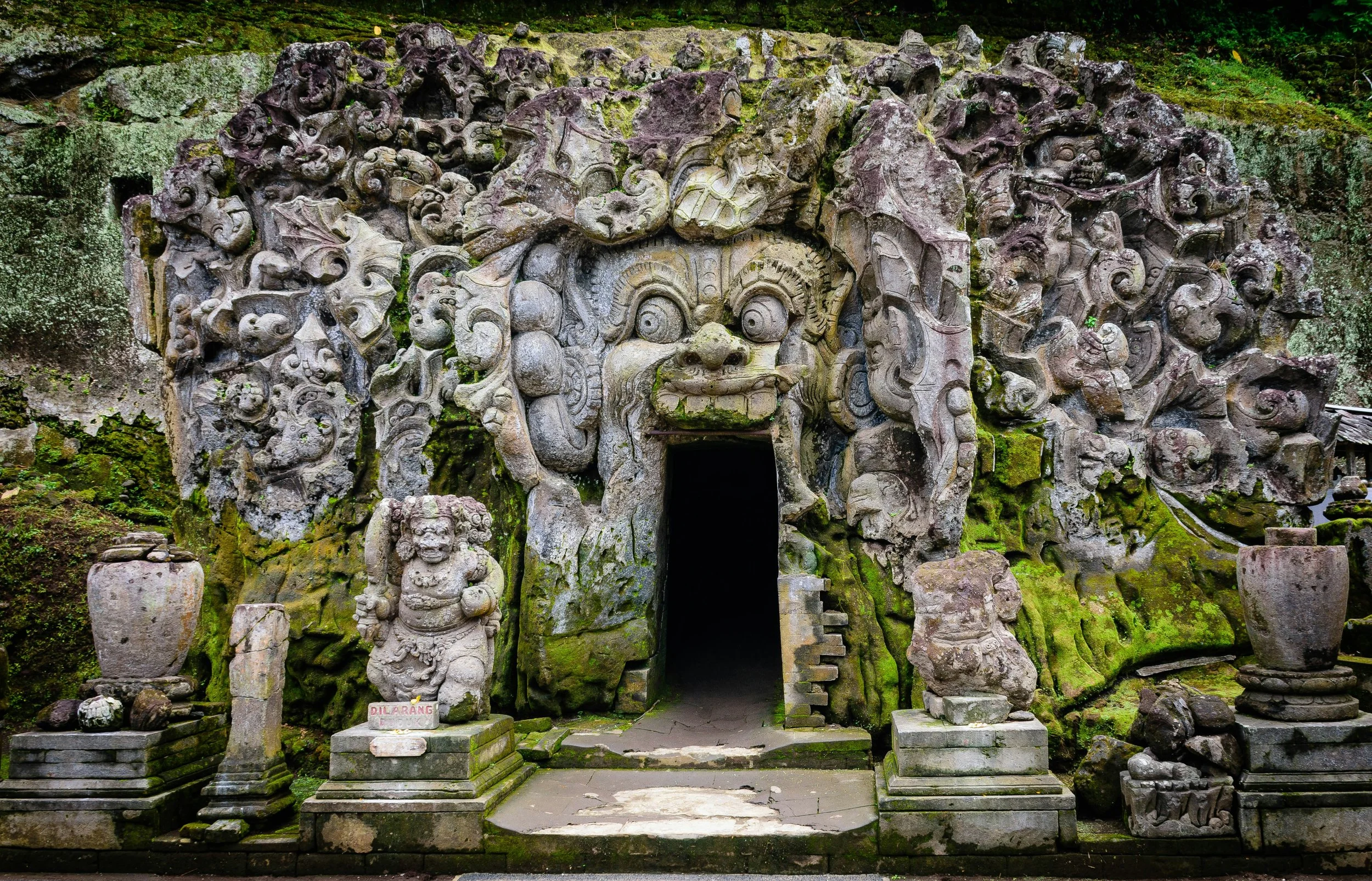 Ancient stone temple entrance decorated with intricate carvings of mythical creatures and faces, moss growing on stones, surrounded by lush greenery.