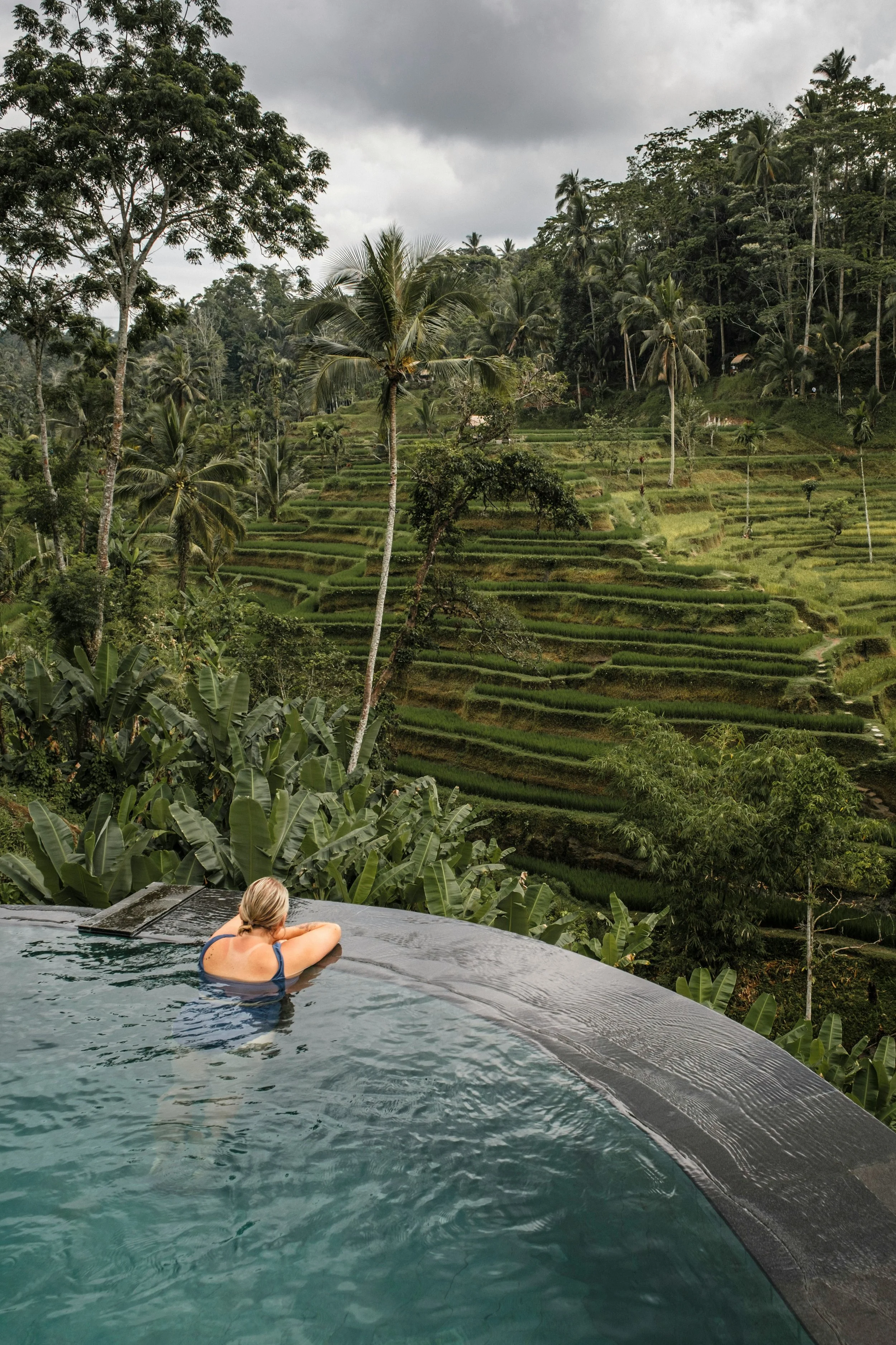 A woman relaxing in a infinity pool overlooking lush green terraced rice fields and tropical trees, with cloudy skies overhead.