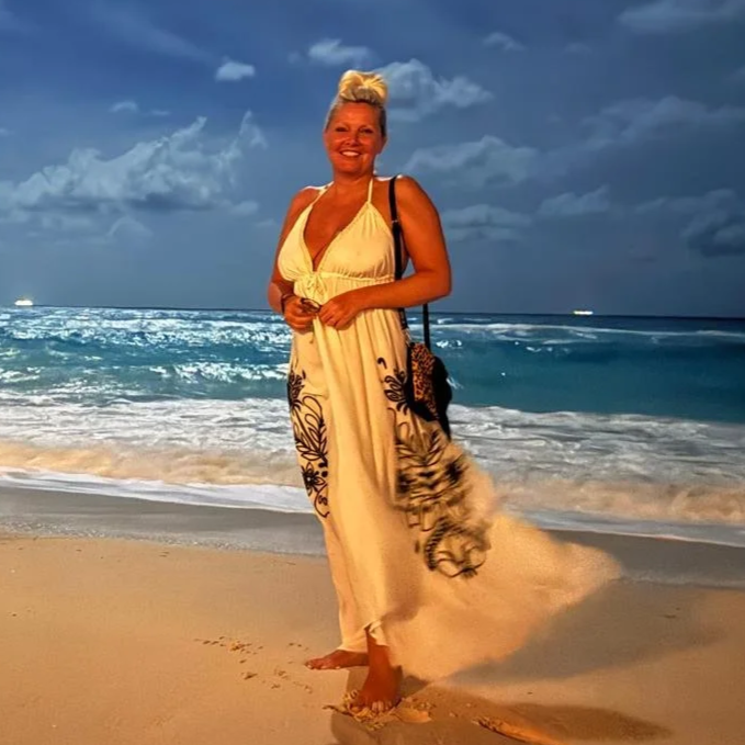 A woman standing barefoot on the beach during sunset, wearing a yellow and black dress, smiling, with the ocean and sky in the background.
