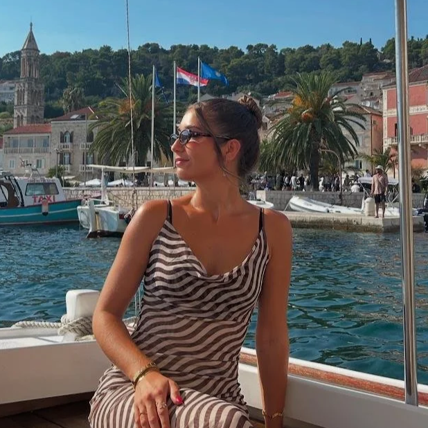 A woman wearing sunglasses and a striped dress sitting on a boat, with a marina with boats and buildings, palm trees, flags, and a hill in the background.