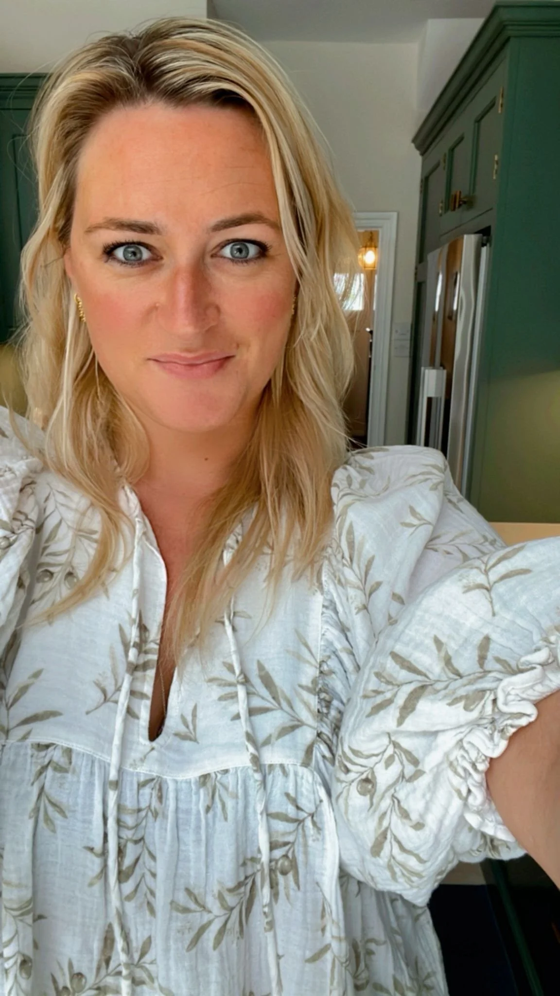 A woman with blonde hair, blue eyes, and earrings, taking a selfie in a room with green cabinets and a refrigerator.