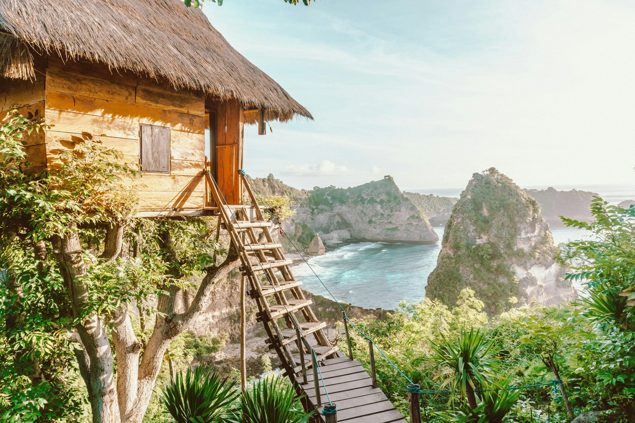 A traditional wooden house with a thatched roof built on stilts near the coast, surrounded by lush green vegetation, with a view of cliffs and the ocean in the background.