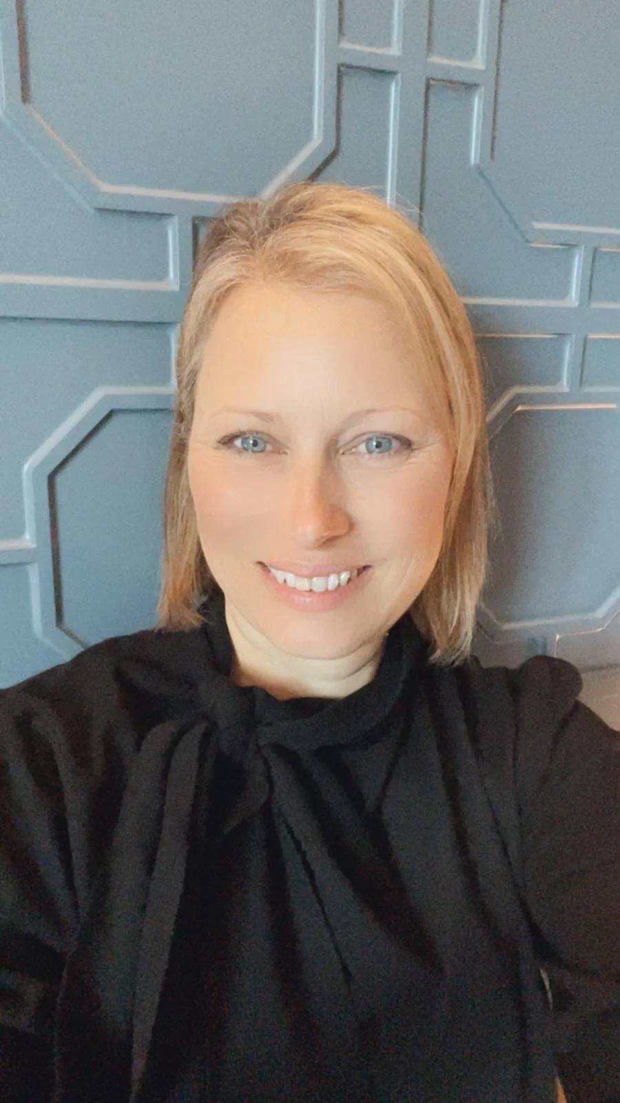 A woman with blonde hair and blue eyes smiling in front of a textured blue wall.