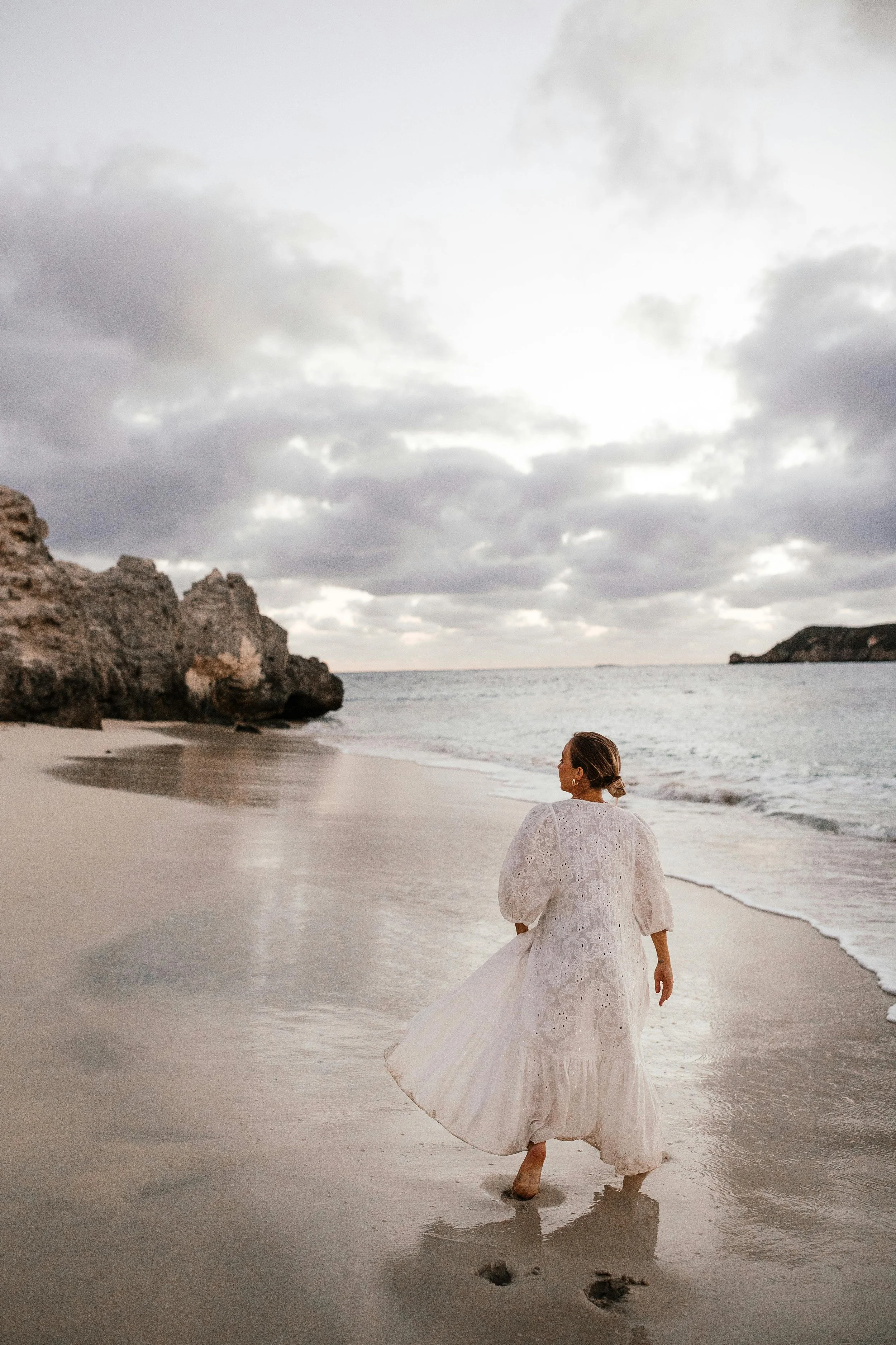 A woman in a white dress walking along a beach with cloudy sky and rocky formations in the background.