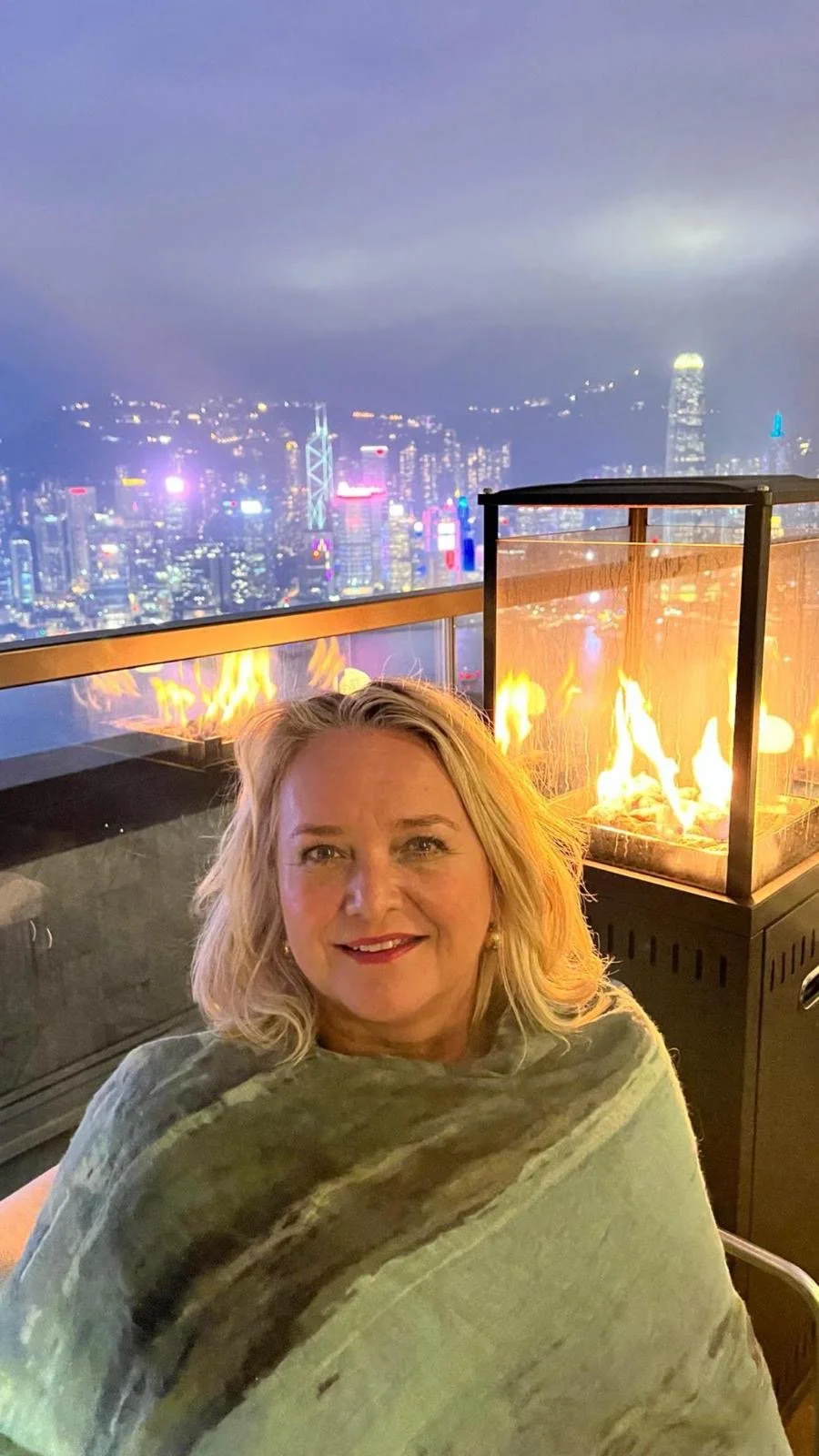 A woman with blonde hair and a shawl smiling at night on a rooftop with a city skyline and a fire feature in the background.
