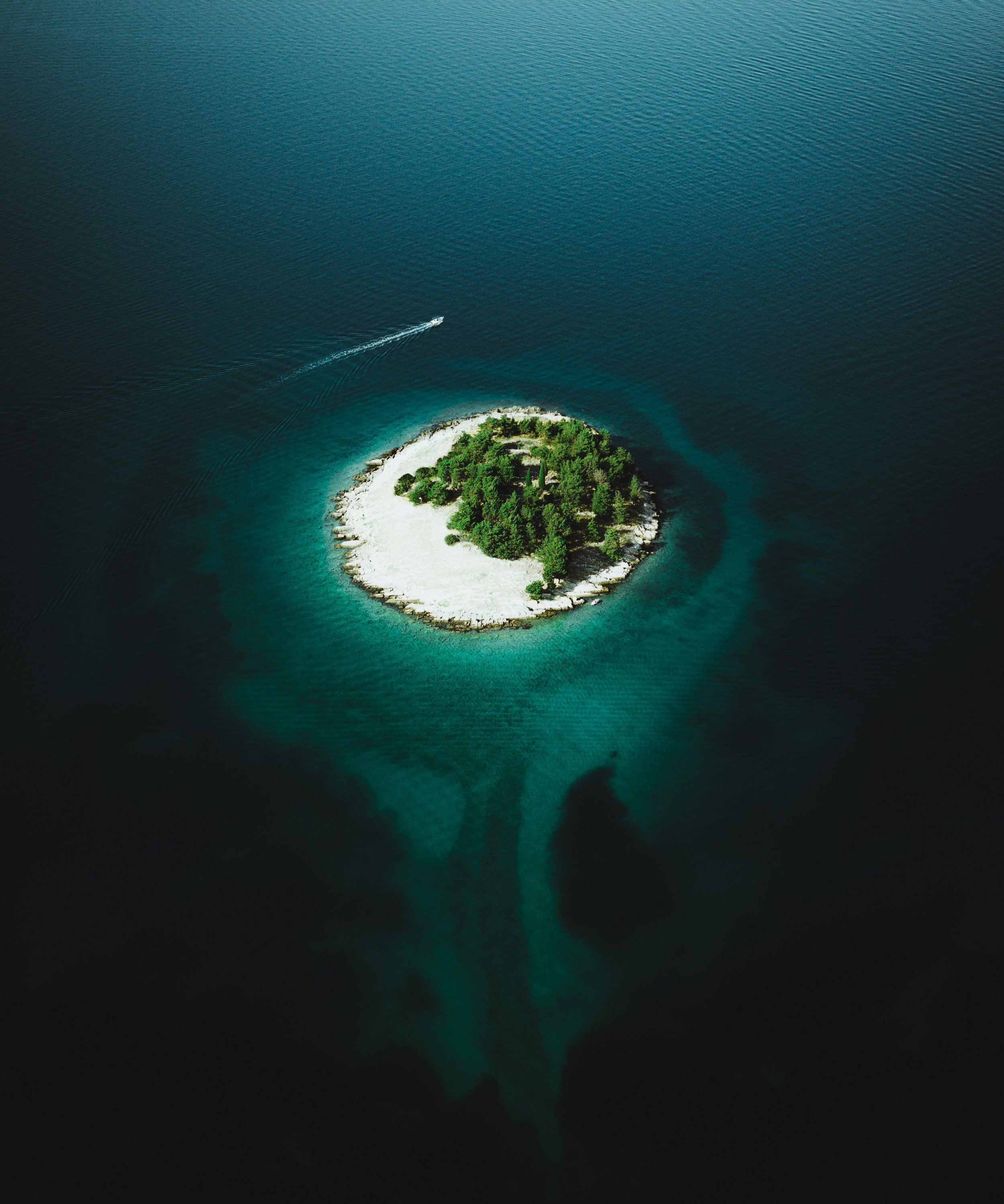 An aerial view of a small, round, sandy island with lush green trees surrounding it in the middle of a deep blue body of water, with a boat creating a wake as it moves away from the island.
