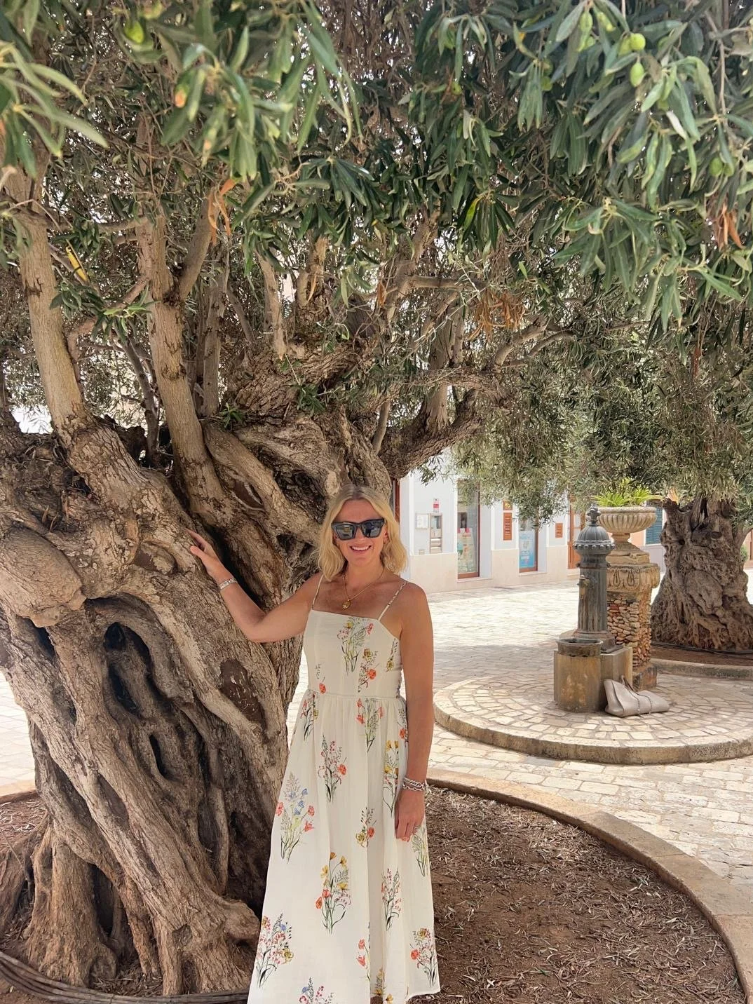 A woman wearing a white floral dress and sunglasses standing next to a large, gnarled tree in a plaza with buildings, a fountain, and shoes on the ground.