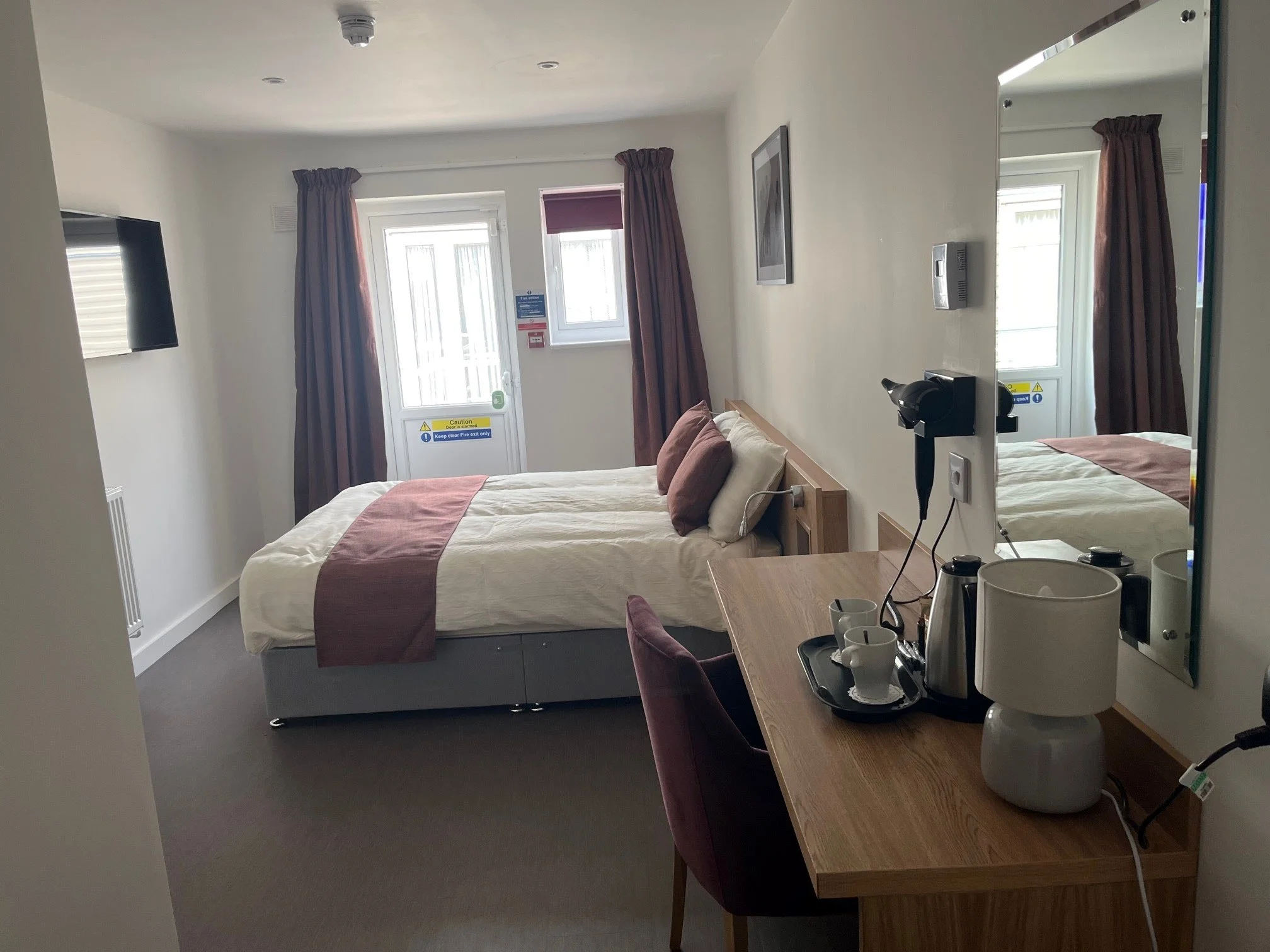 Double bed in a hotel bedroom with cream bedding and maroon curtains.