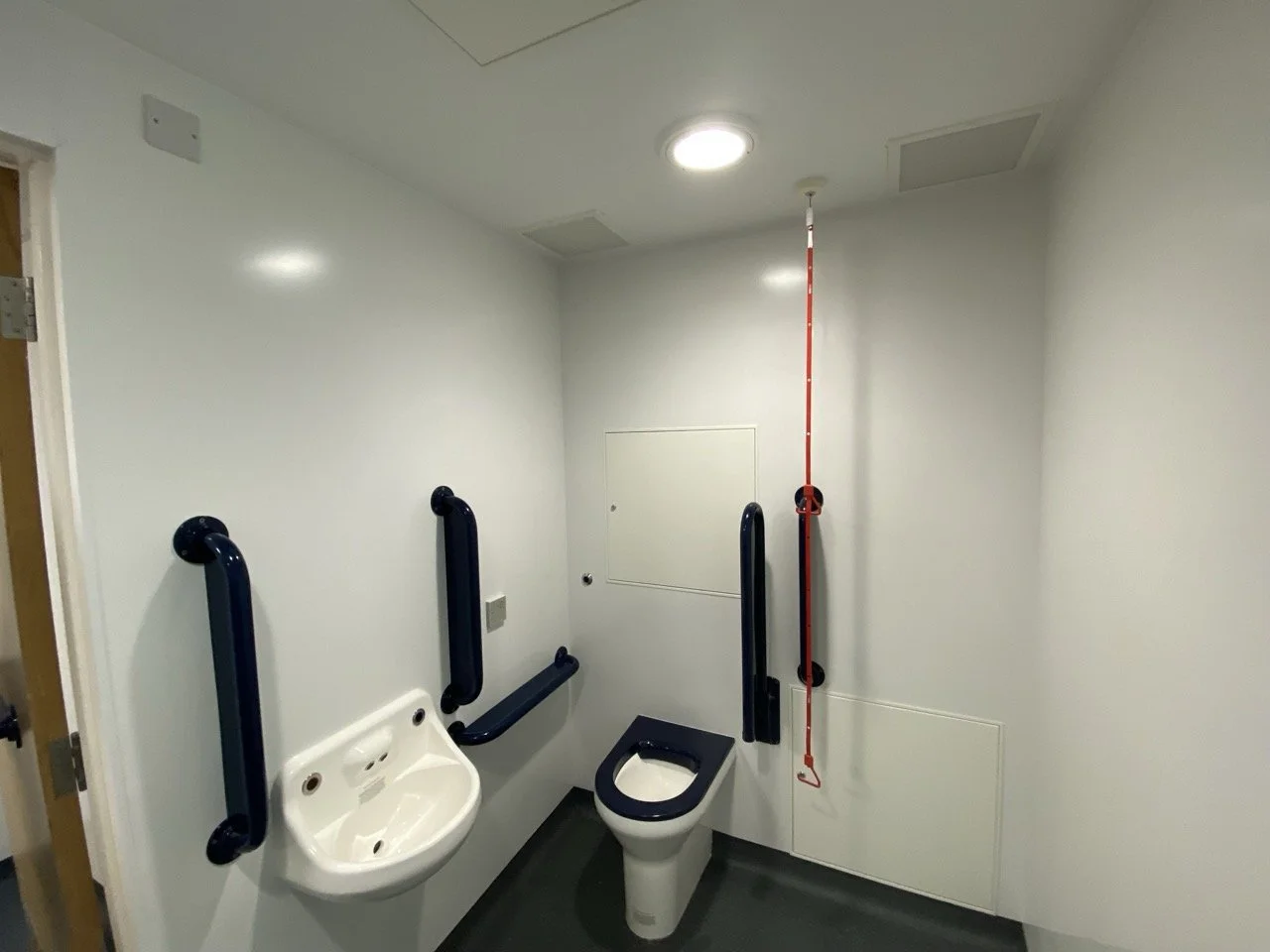 White-walled disabled toilet with black chrome handles on the wall and a red emergency pull.
