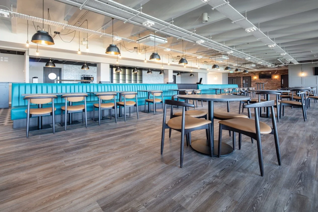 Large seating area in a bar with padded electric blue benches and fawn-cushioned chairs.