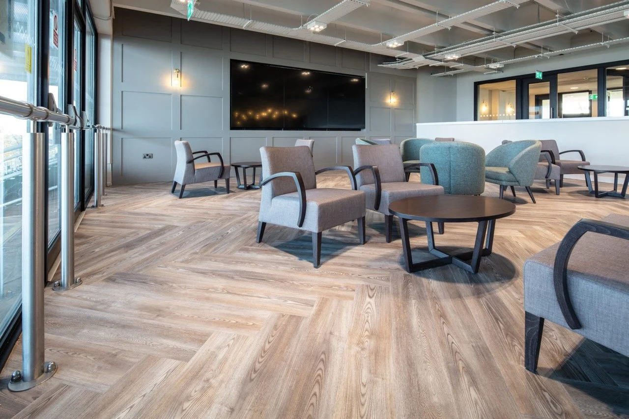 Hospitality area over looking a racecourse with a large tv screen set in a grey panelled wall. Pale armchairs and formal sofas are dotted around the room.