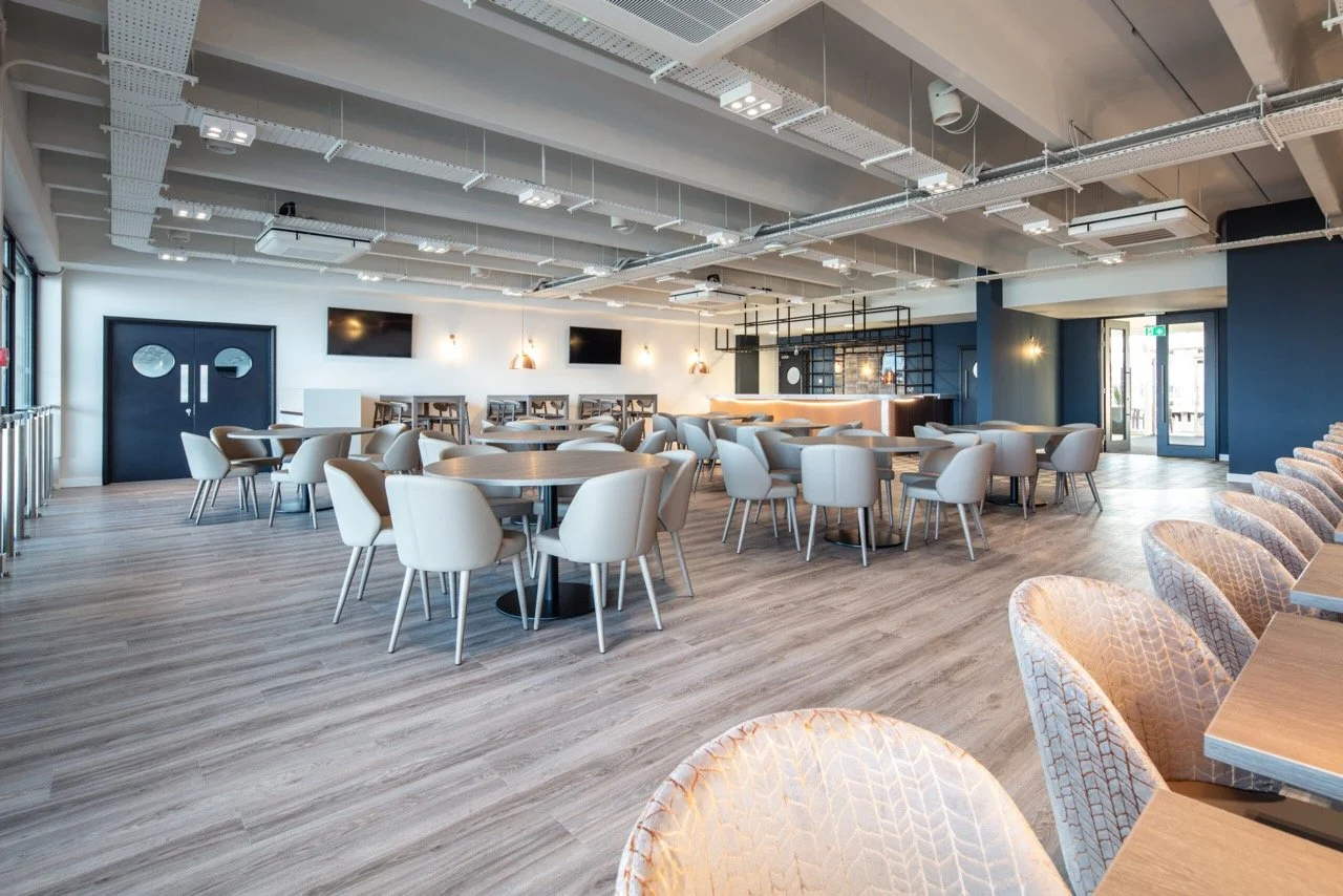 Hospitality area over looking a racecourse with a large tv screen set in a grey panelled wall. Pale armchairs and formal sofas are dotted around the room.