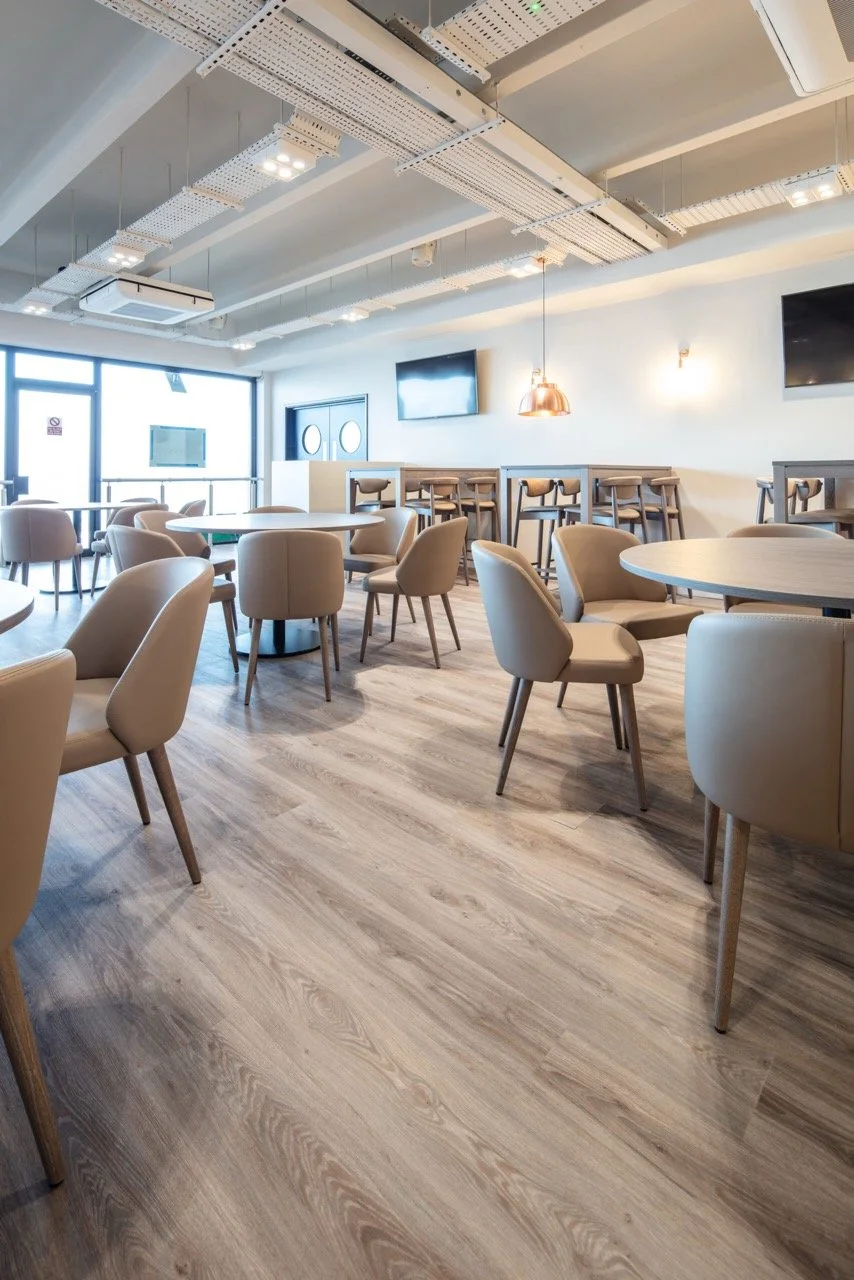 Hospitality area over looking a racecourse with a large tv screen set in a grey panelled wall. Pale armchairs and formal sofas are dotted around the room.