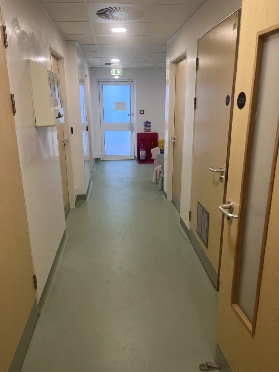 An empty health centre corridor with sage green Lino flooring and pale pine doors leading off into individual clinician rooms.