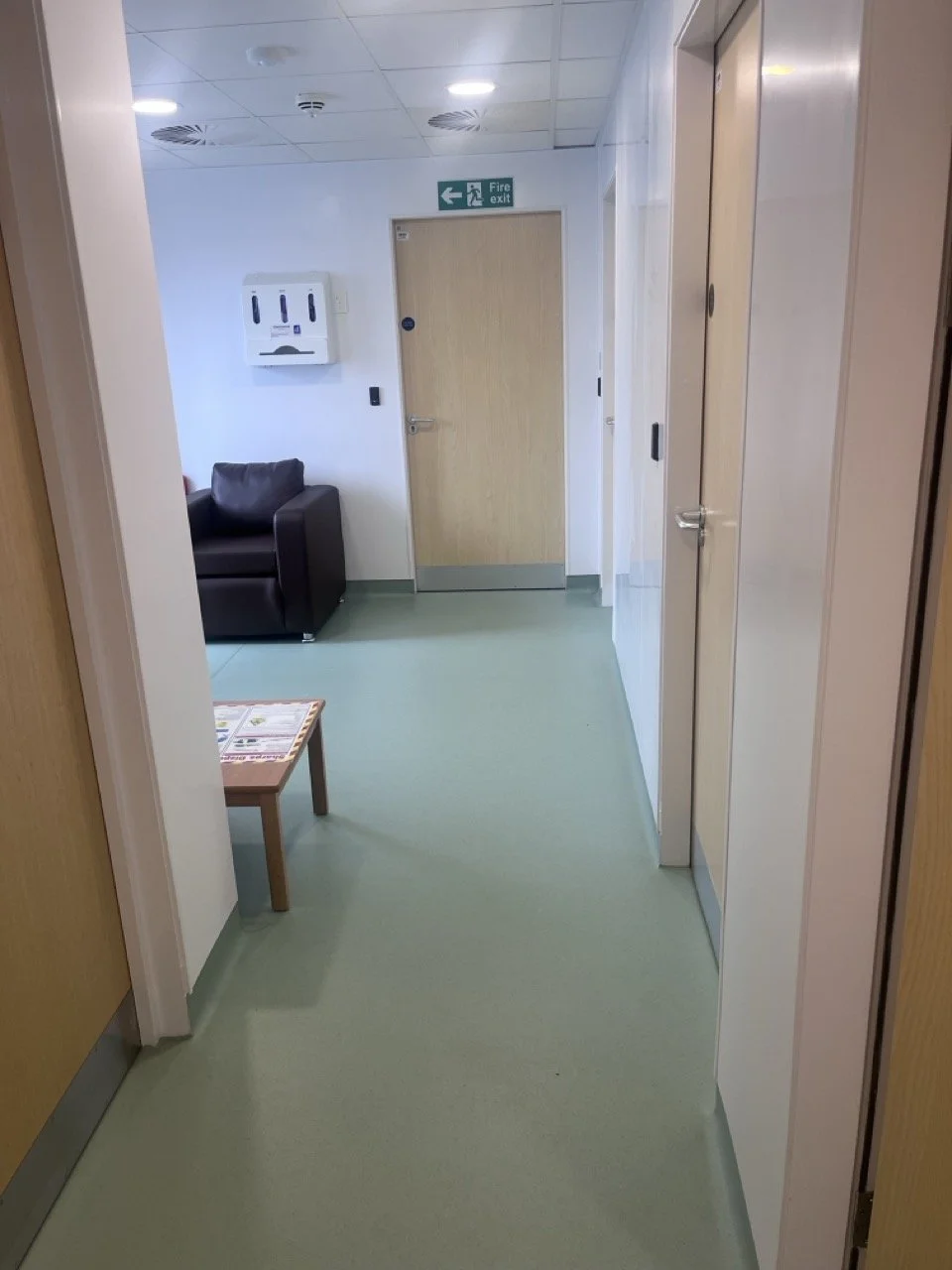 Looking into a healthcare waiting area with sage green Lino floor - a single brown leather chair can be seen underneath a hygiene glove dispenser.