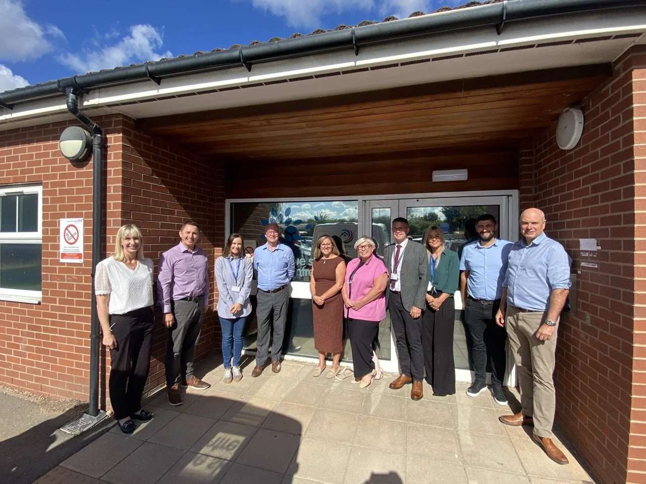 A group of eleven people standing outside a brick healthcare building, smiling for a photo on a sunny day.