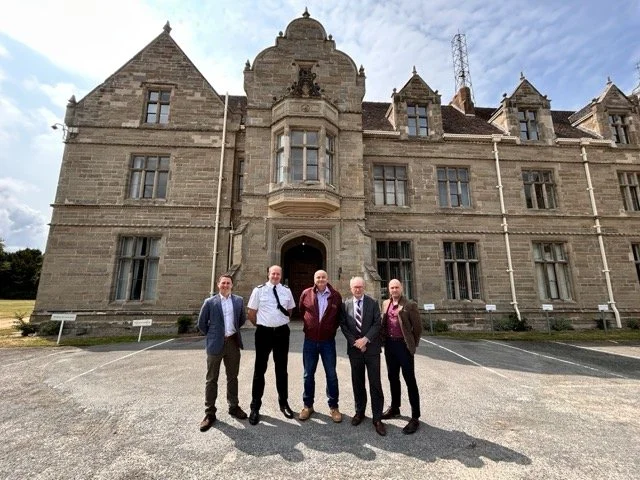 Five men stand in front of an old stone two-story Scottish building. Four wear shirts, and one wears police uniform.