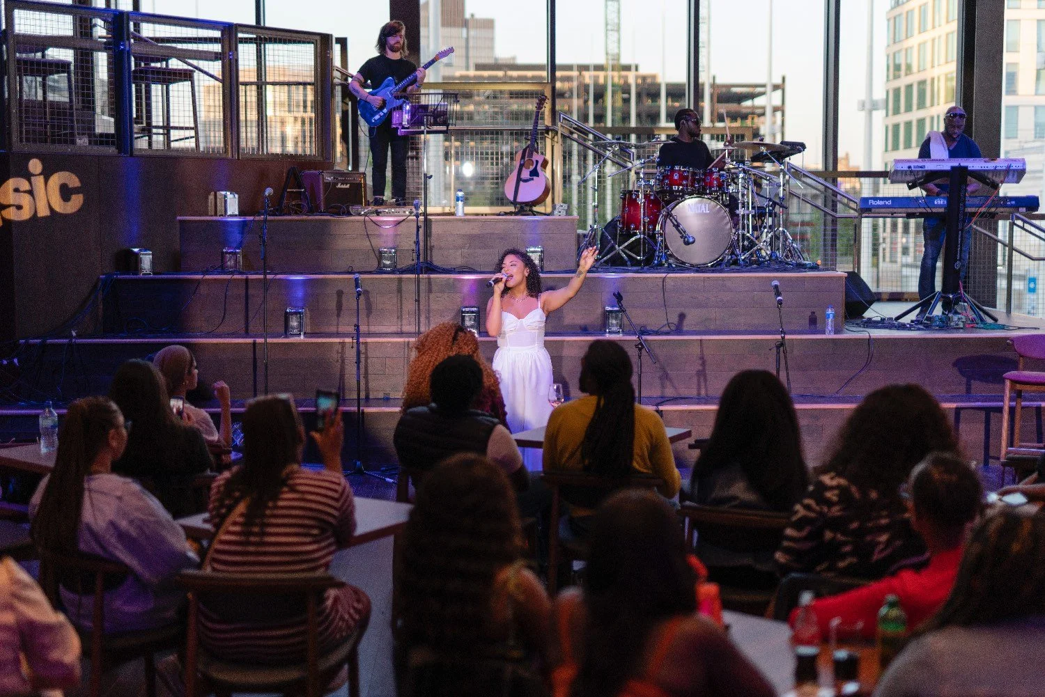 A female wearing a white dress sings into a microphone at an entertainment  venue - in front of her are people sitting at tables and chairs.