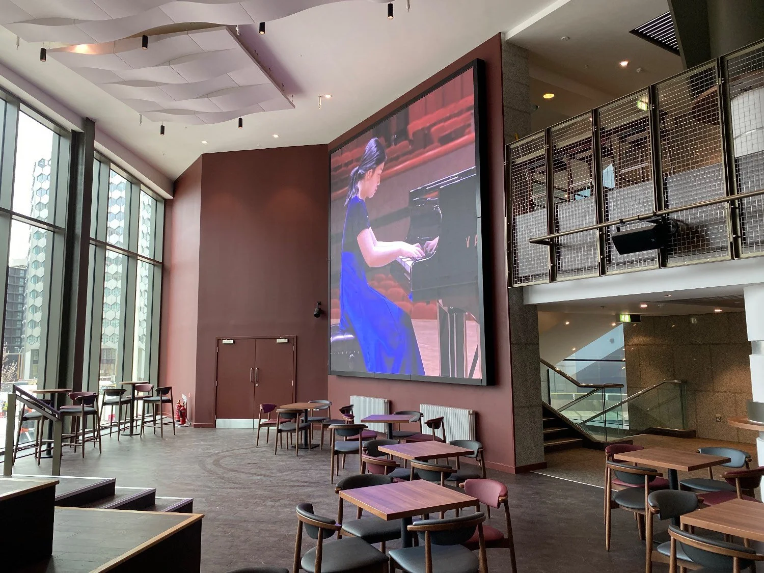 A large, ceiling-height screen shows a female performing at a piano in a midnight blue dress. The screen is in a maroon-walled room with tables and chairs.