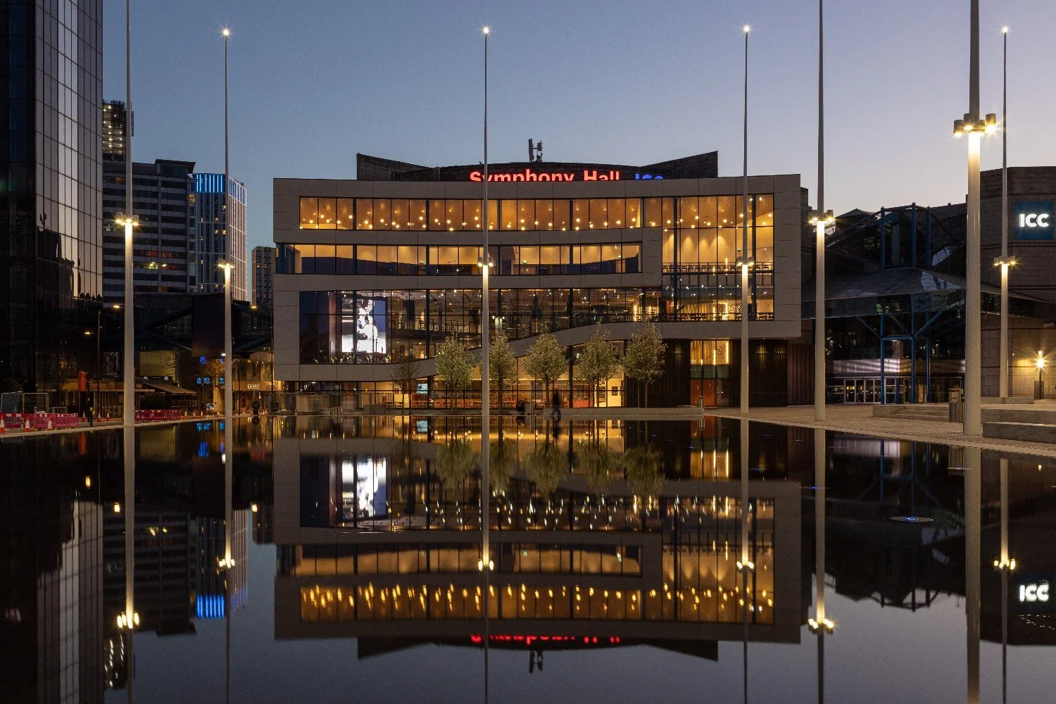A large modern building with flag poles is lit-up at night. It sits behind a manmade lake and is reflected in the water.