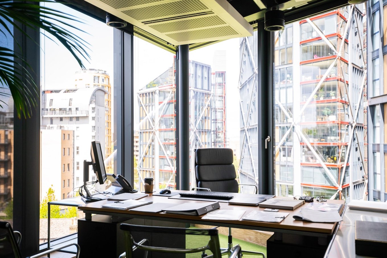 Glass walled, light-filled office with black framed windows and a large black desk with a screen, telephone and coffee cup.