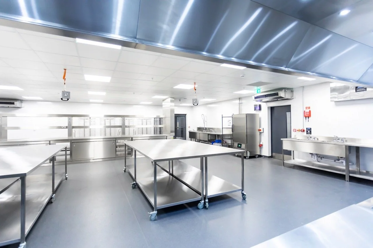 Glistening silver commercial kitchen with preparation tables and large silver fridges.