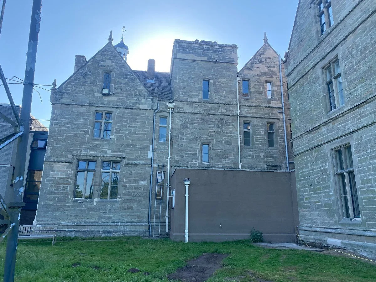 An old, large stone building with two stories on a sunny day. There is an area cornered-off by metal fencing suggesting building work is being completed.