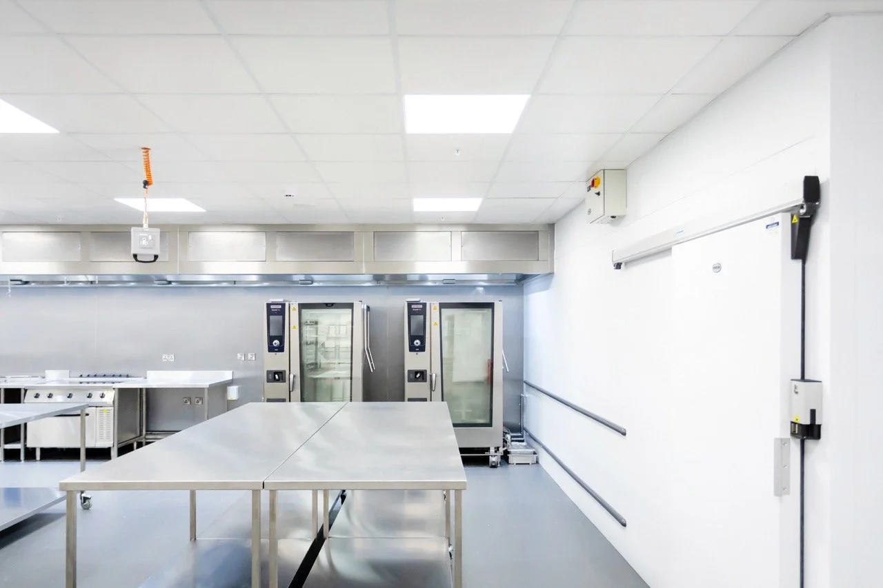 Glistening silver commercial kitchen with preparation tables and large silver fridges.
