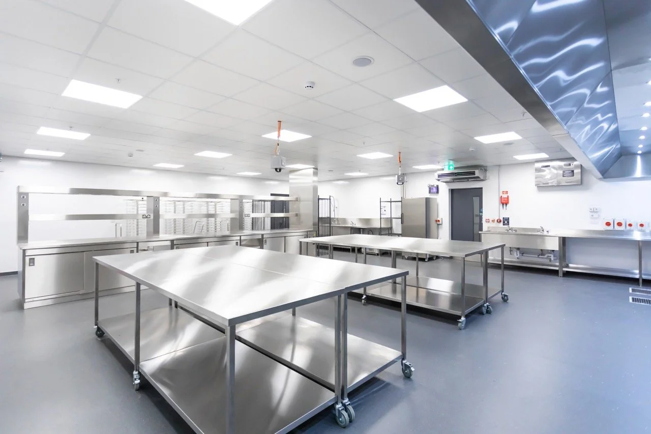 Glistening silver commercial kitchen with preparation tables and large silver fridges.