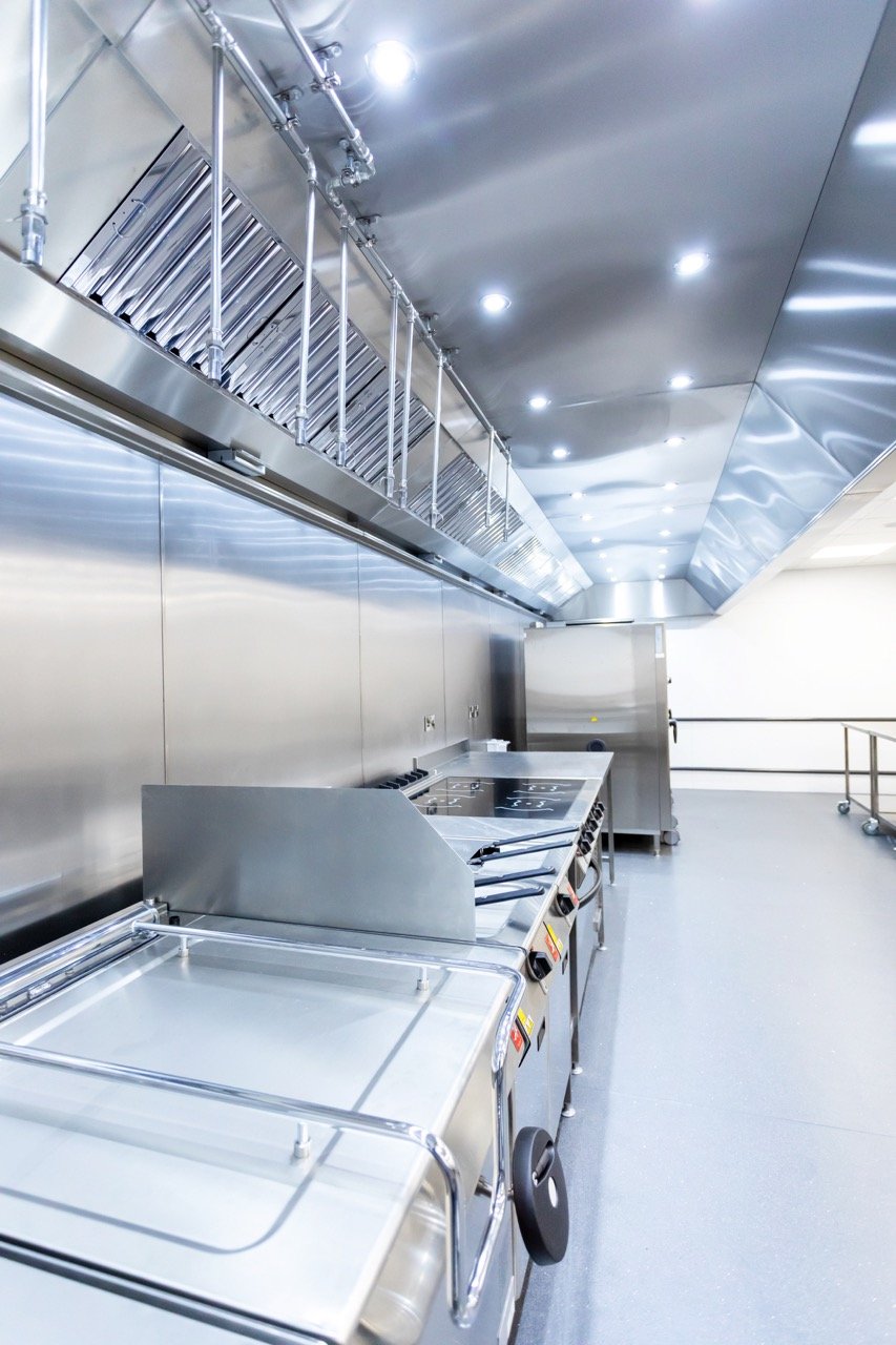Glistening silver commercial kitchen with rows of shiny silver kitchen units and bright ceiling lights glowing.