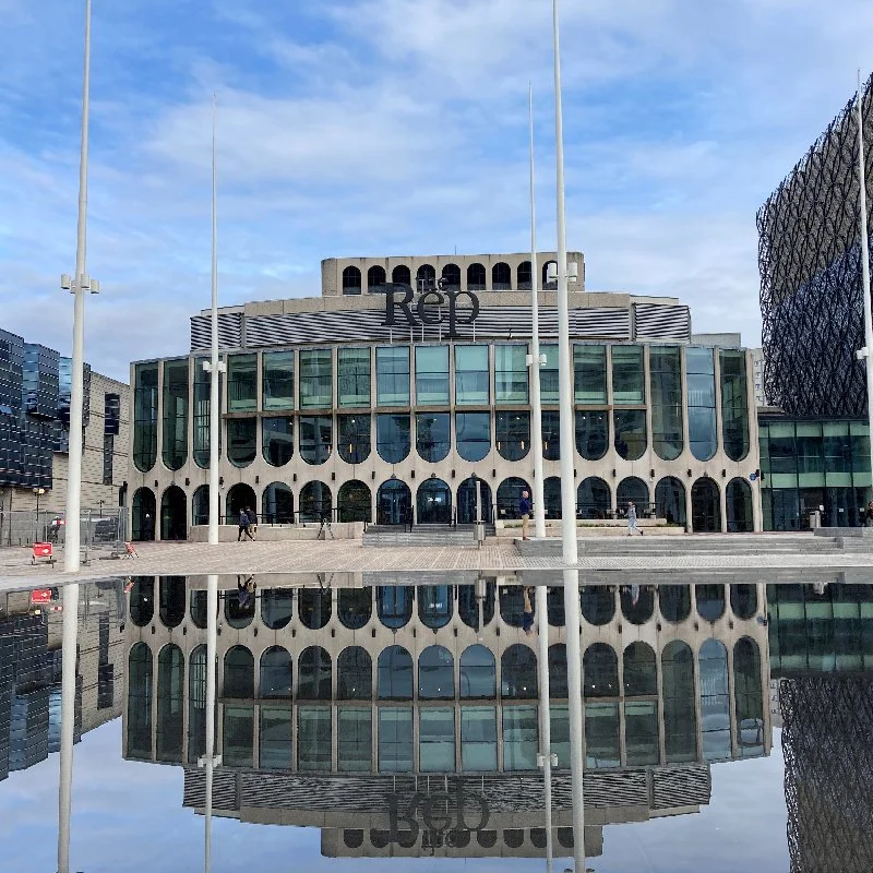 building with a unique architectural design, featuring large glass windows and rounded arches. There are flagpoles in front of the building, and the reflection of the building and flagpoles can be seen in a water feature or puddle in the foreground. A few people are walking near the entrance.