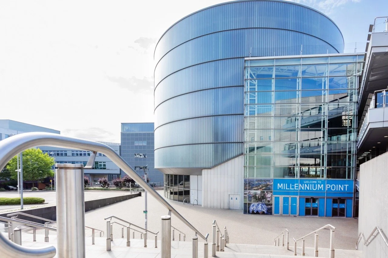 Large, modern, circular building with panelled glass around the outside, set in an urban area with wide steps leading down to the entrance..