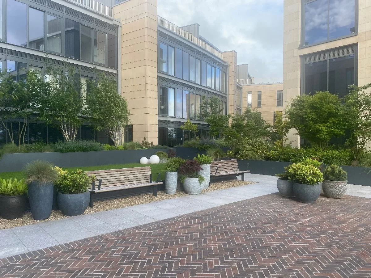 Urban patio with benches, planters filled with greenery, and modern apartment buildings in the background.