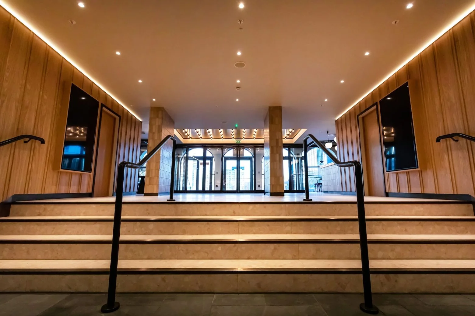 Large, empty leisure venue entrance with wide cream marble steps with black trips and two large black handrails leading up to three double class doors.